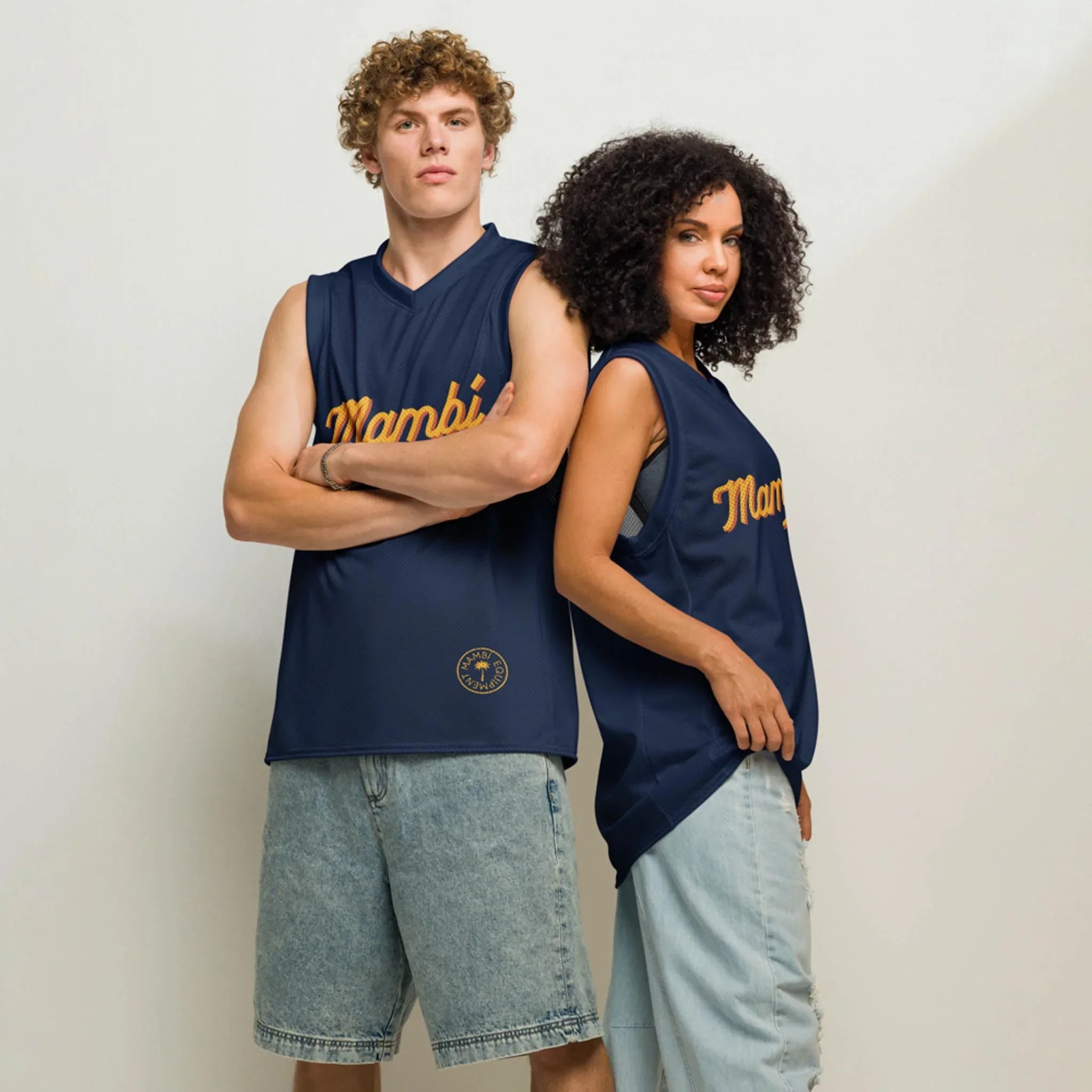 Two people stand back to back in Printful’s Yordan Premium Basketball Jersey | Unisex, featuring navy blue fabric with Mamba in yellow. One pairs it with denim shorts, the other with a skirt—showcasing versatile style and comfort.