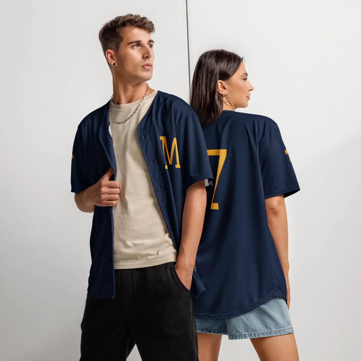 A young man and woman wear Printfuls unisex Zoilo Classic Baseball Jersey in navy; he faces forward showing the M on the front, while she faces away revealing the number 7 on the back.
