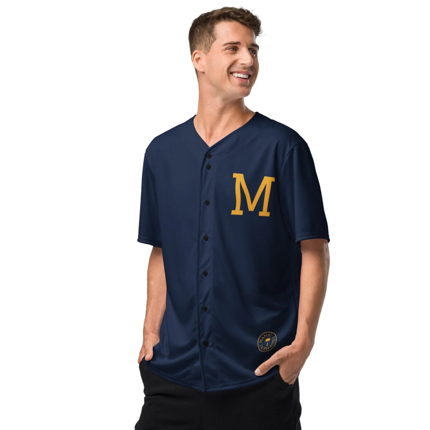 A smiling man stands hands in pockets, wearing the Printful Zoilo Classic Baseball Jersey | Unisex in navy blue, featuring a large yellow M on the chest and a round patch near the hem.