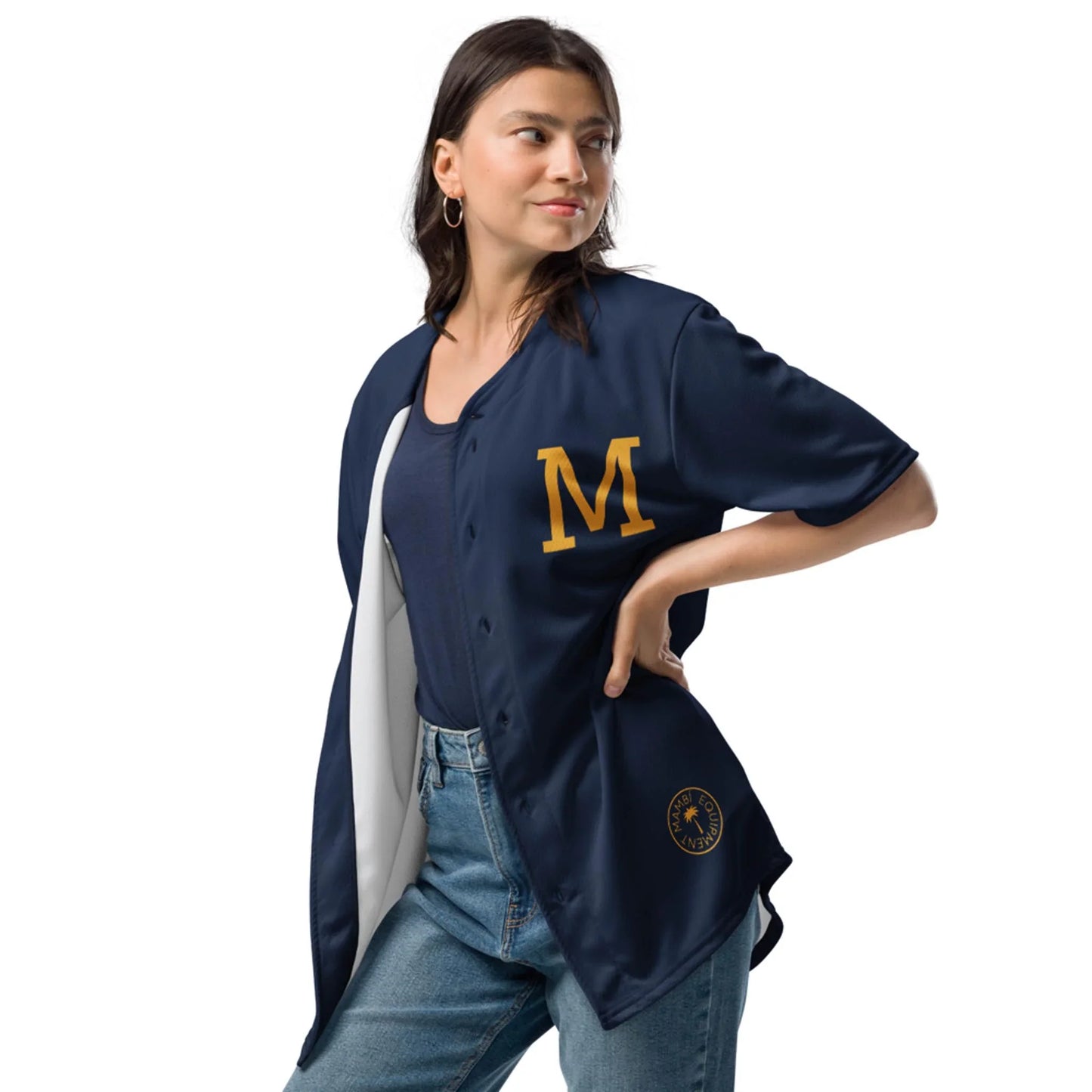 A woman stands with one hand on her hip, wearing the Printful Zoilo Classic Baseball Jersey | Unisex, featuring a large yellow M on the chest. She pairs it with blue jeans and hoop earrings, looking off to the side with a slight smile.