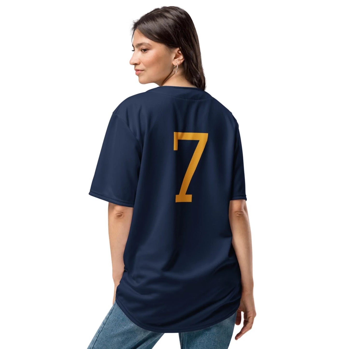A woman with long dark hair wears the Zoilo Classic Baseball Jersey by Printful, featuring a large yellow number 7 on the back. She pairs this unisex navy blue jersey with blue jeans and looks over her shoulder in a relaxed pose.
