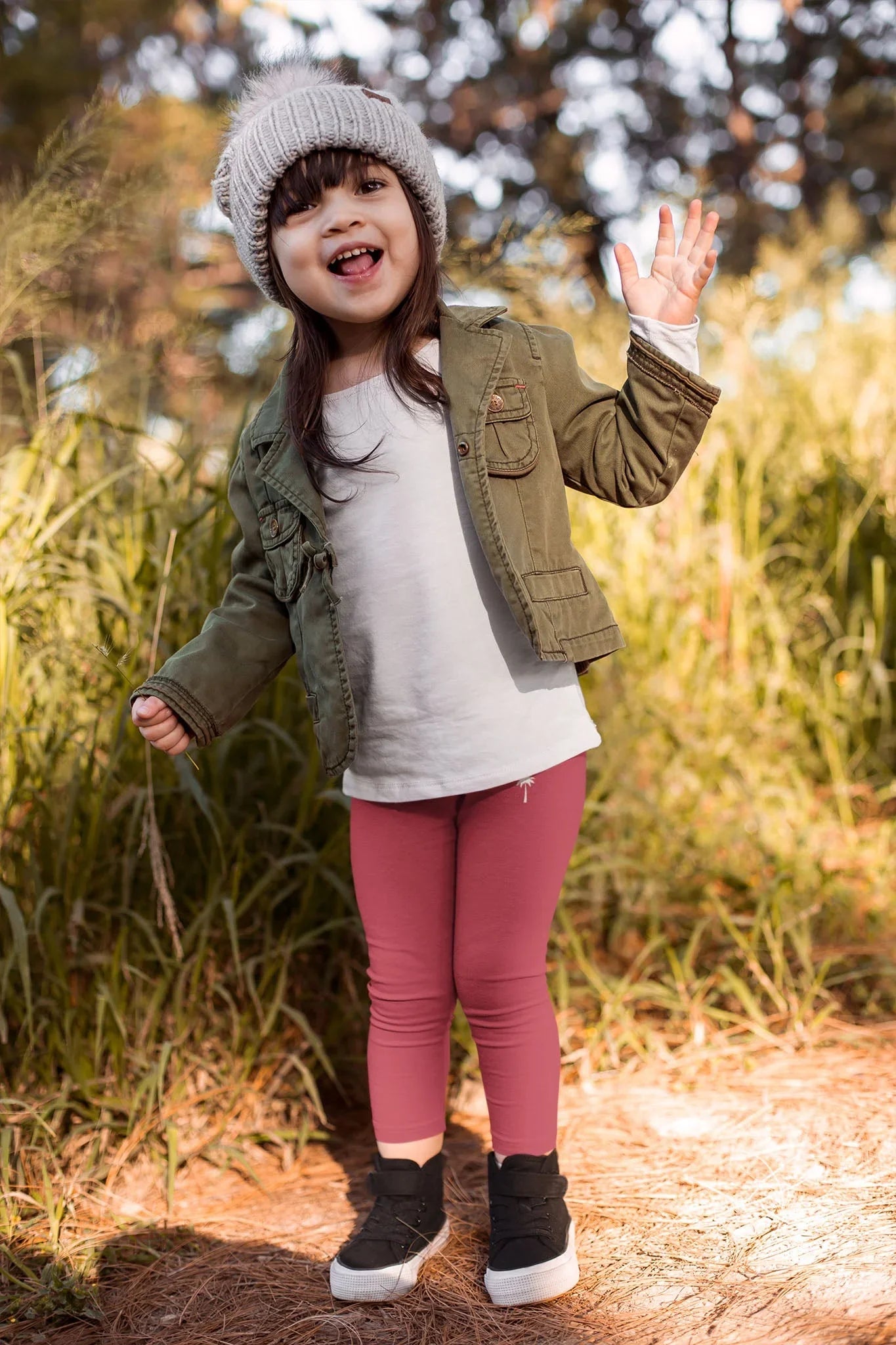 Smiling child in knit beanie, green jacket, pink leggings outdoors in nature