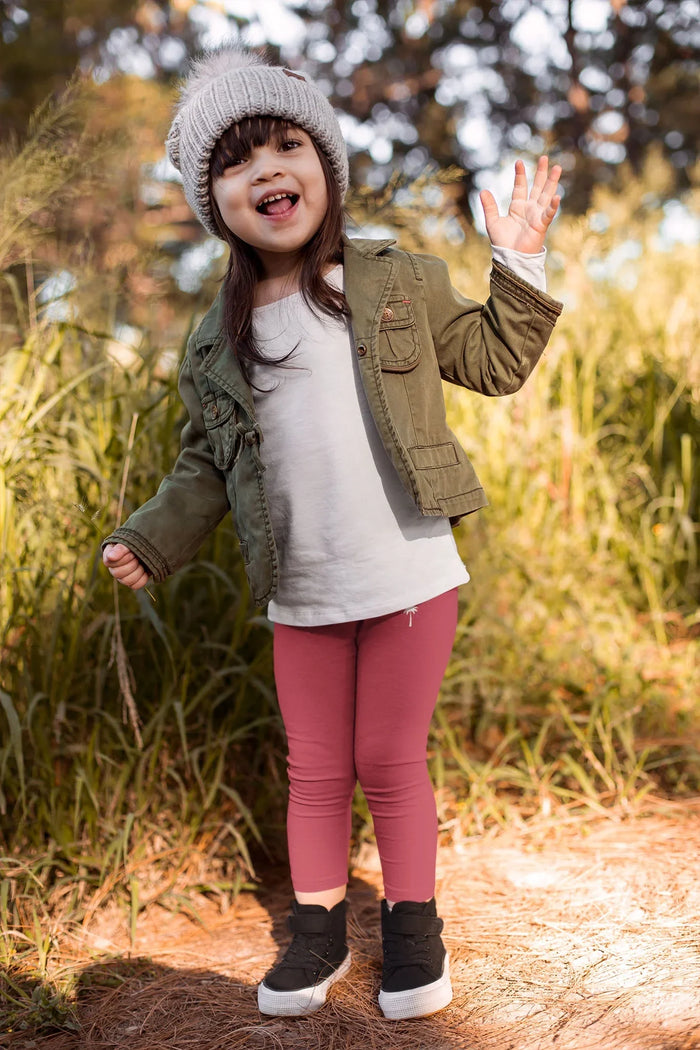 Smiling child in knit beanie, green jacket, pink leggings outdoors in nature