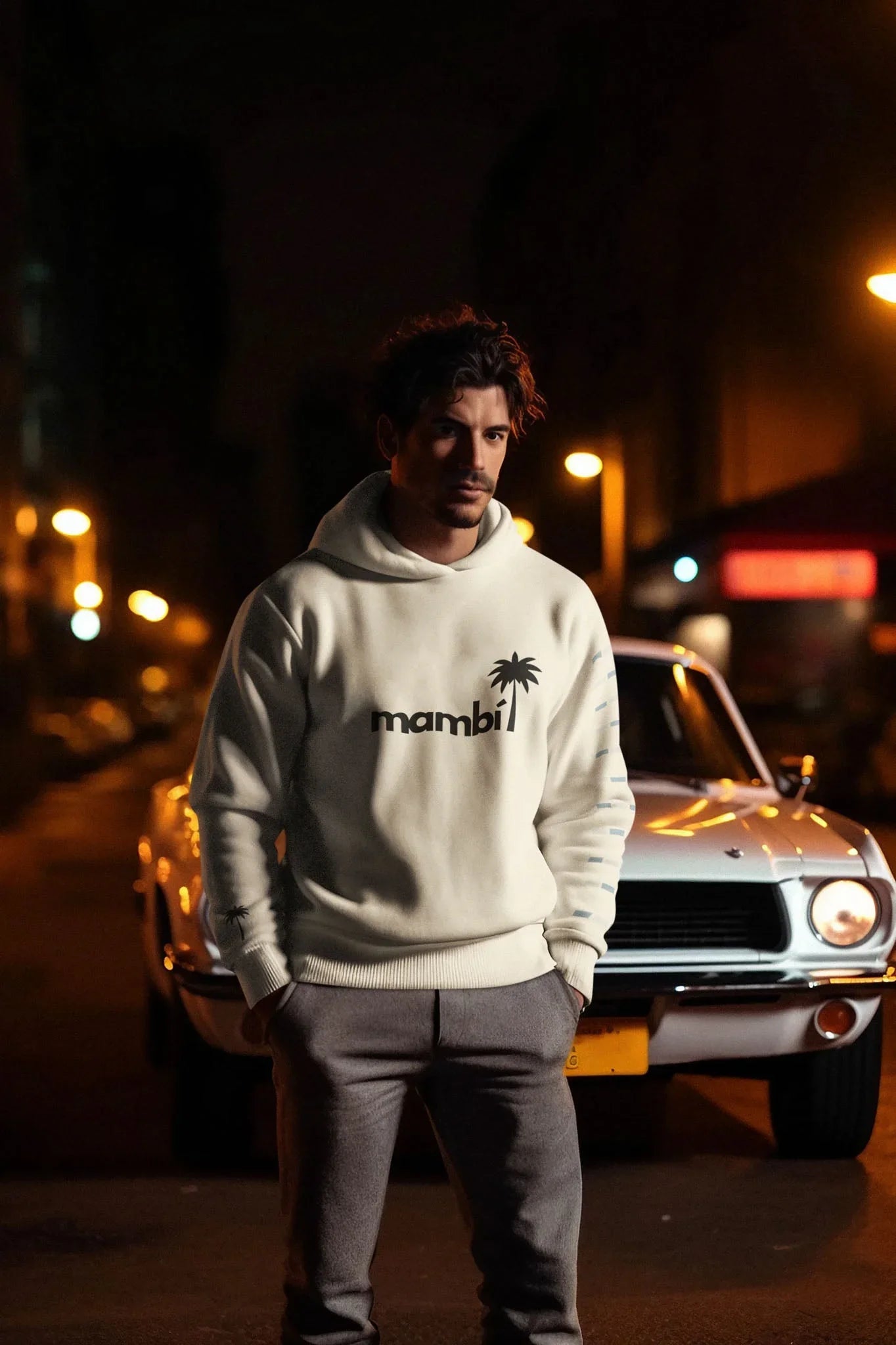 A man in a white mambi hoodie and gray pants stands with hands in pockets in front of a vintage white car on a city street at night, illuminated by streetlights.