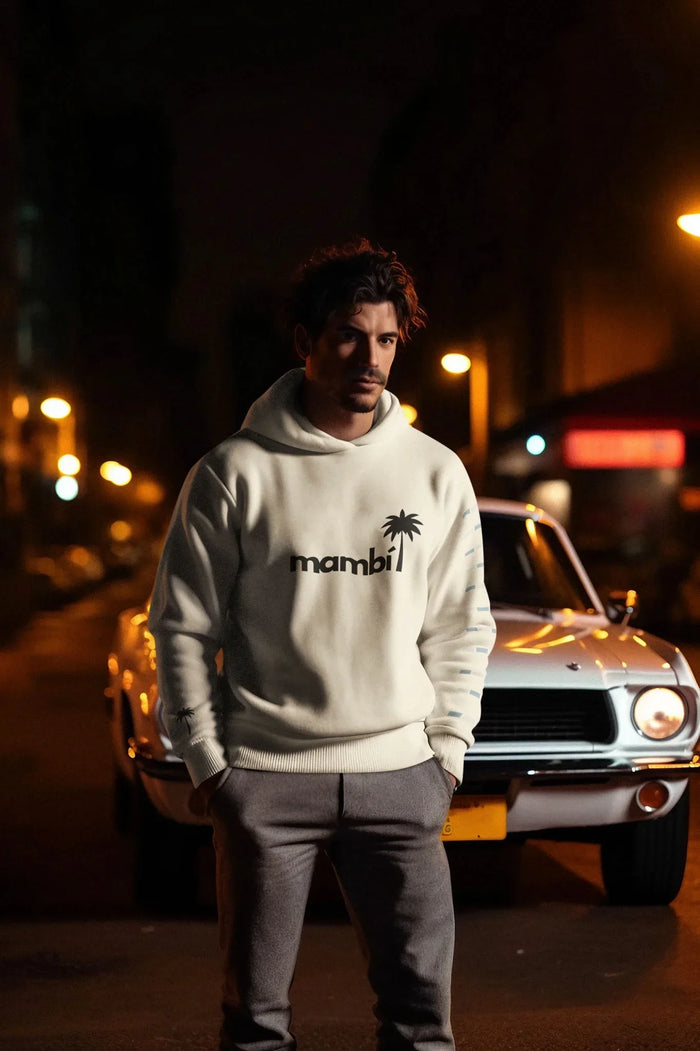 A man in a white mambi hoodie and gray pants stands with hands in pockets in front of a vintage white car on a city street at night, illuminated by streetlights.