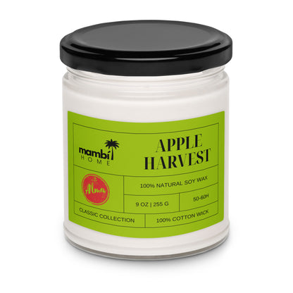 A white candle in a glass jar with a black lid. The green label reads APPLE HARVEST, mambi HOME, and highlights this eco-friendly candle made from 100% natural soy wax, 9 oz size, 50-60h burn time, and a 100% cotton wick.