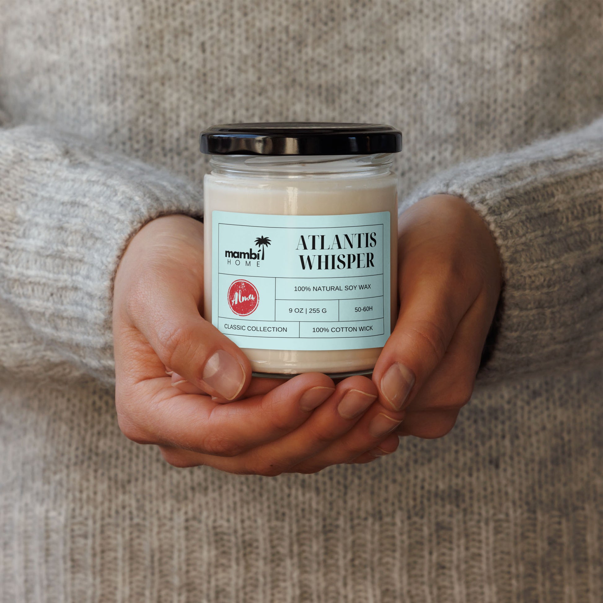 A person wearing a light grey sweater holds a glass jar candle labeled ATLANTIS WHISPER, made from 100% natural soy wax and 100% cotton wick—perfect for those who love natural soy wax candles.