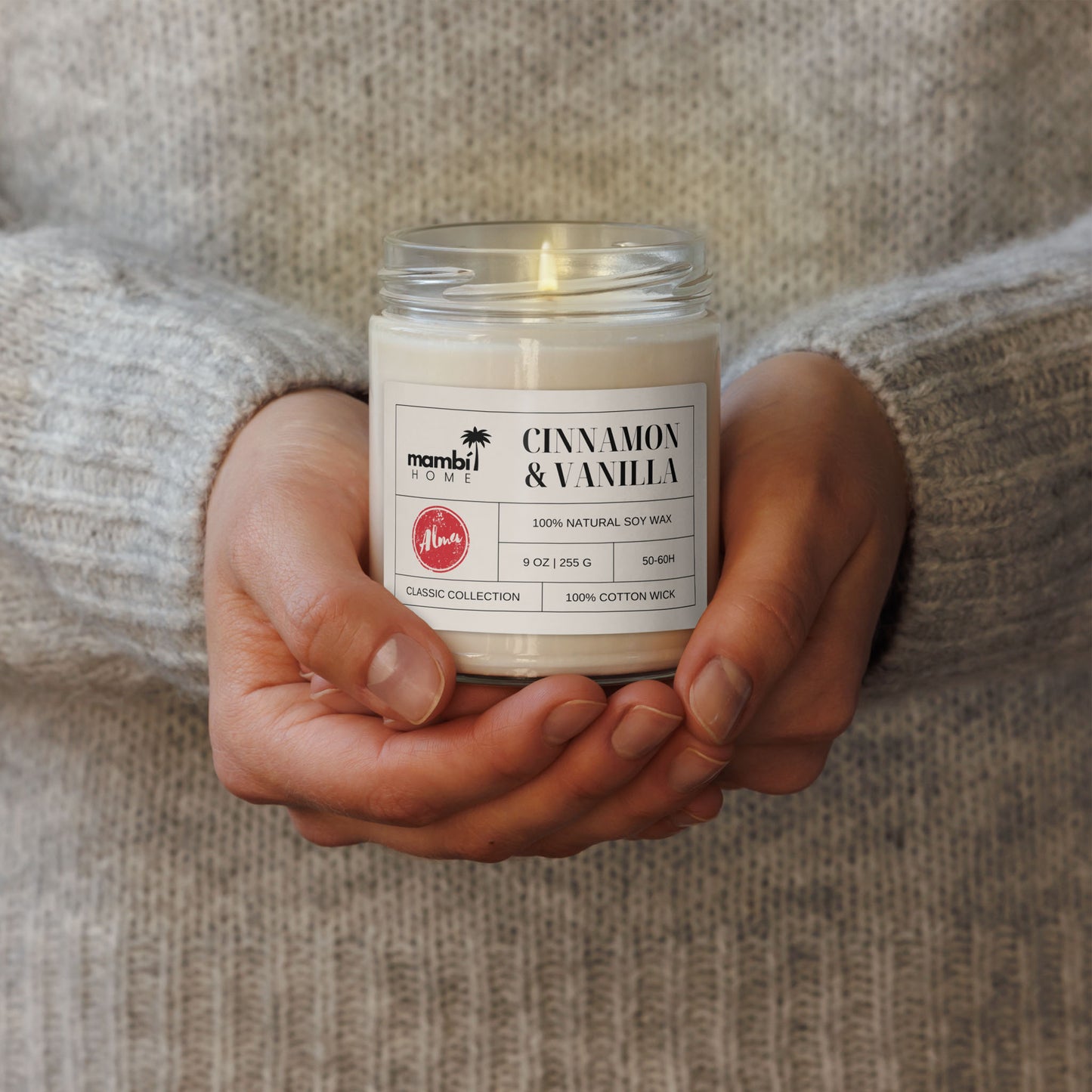A person in a cozy, light gray sweater holds a lit jar candle labeled Cinnamon & Vanilla with both hands. These scented soy candles are made of 100% natural soy wax for a clean burn and long-lasting fragrance.
