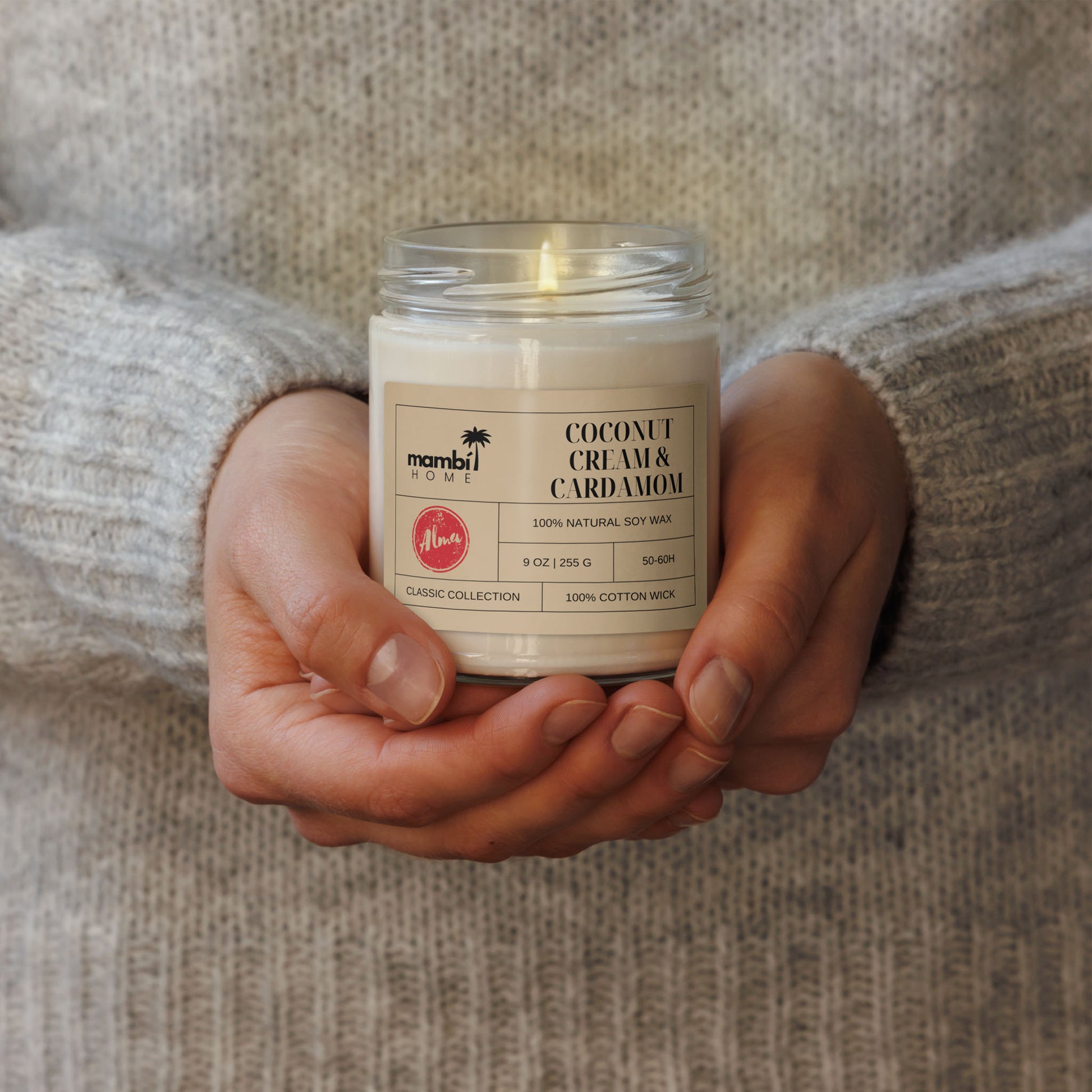 A person in a cozy, light gray sweater holds a glass jar candle labeled Coconut Cream & Cardamom, made from natural soy wax. This Classic Collection piece highlights the appeal of eco-friendly candles for any relaxing space.