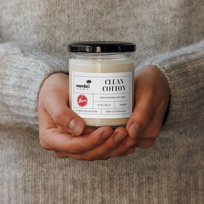 A person wearing a light grey, knit sweater holds a glass jar candle labeled Clean Cotton, made with 100% natural soy wax. This cozy scene highlights the appeal of eco-friendly candles for a fresh and inviting home atmosphere.
