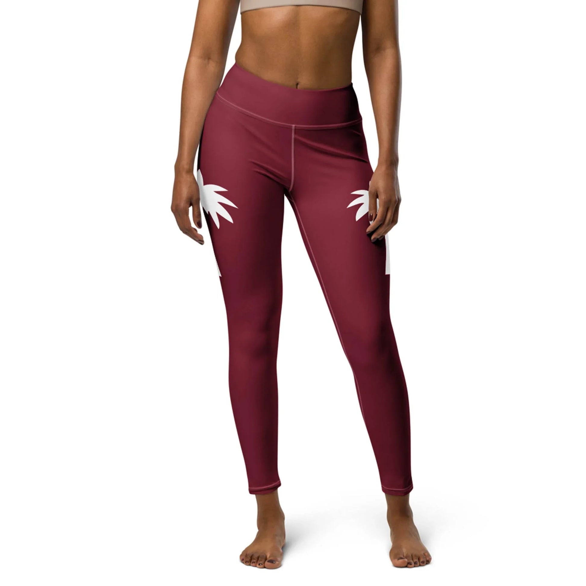 A woman models Printful’s Alpine High-Rise Yoga Leggings in maroon with white palm tree designs, paired with a beige sports bra, embracing athleisure while standing barefoot against a white background.