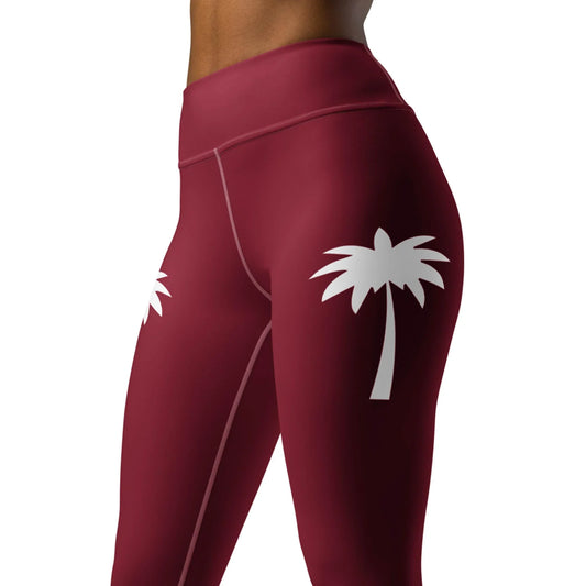 A person models Printfuls Alpine High-Rise Yoga Leggings for women in maroon, featuring white palm tree graphics on the hips and four-way stretch, shown waist to mid-thigh against a white background for a stylish athleisure look.