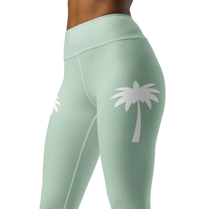 A woman models Printfuls Alpine High-Rise Yoga Leggings for women in light green with white palm tree graphics on the outer thighs. The cropped image highlights her waist and legs, emphasizing the stylish athleisure design.