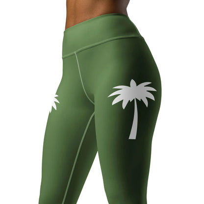 A woman models Printfuls Alpine High-Rise Yoga Leggings in olive green, made with four-way stretch fabric and white palm tree graphics on the thighs. The cropped image highlights the torso and upper legs against a plain backdrop.