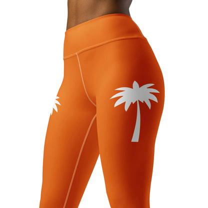 A model wears Printfuls Alpine High-Rise Yoga Leggings for women in bright orange with white palm tree graphics on the upper thighs, shown waist to mid-thigh against a plain background, offering a vibrant athleisure style.