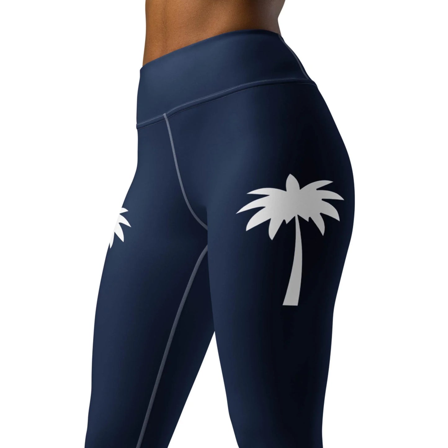 A woman models Printfuls Alpine High-Rise Yoga Leggings in navy blue with a white palm tree graphic on the outer thigh. The cropped image highlights the stylish four-way stretch fabric, perfect for an athleisure look.