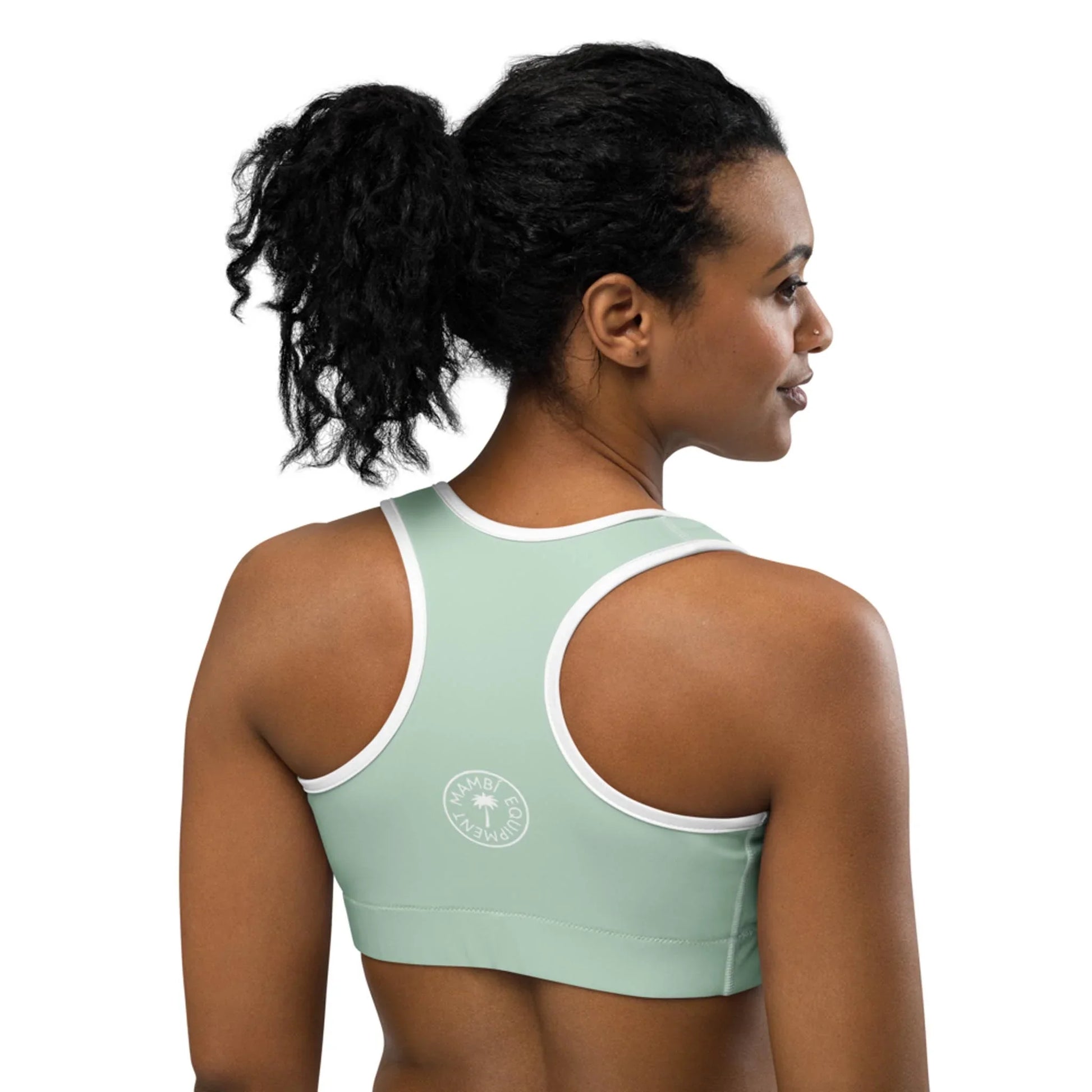 A woman with curly black hair in a ponytail wears a light green Mambí sports bra with white trim. She is facing away, highlighting the back of the bra and its circular white logo on the left side.