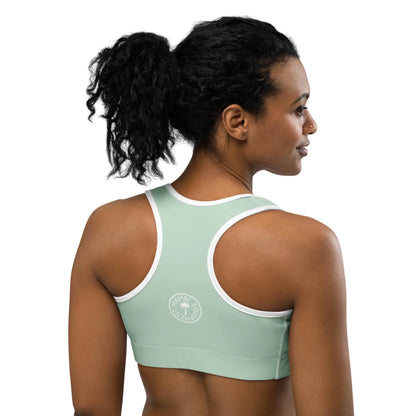 A woman with curly black hair in a ponytail wears a light green Mambí sports bra with white trim. She is facing away, highlighting the back of the bra and its circular white logo on the left side.