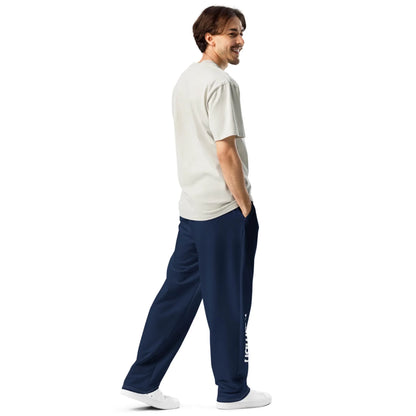 A man stands smiling, facing sideways and looking back. He wears a light gray Mambí t-shirt, navy blue sweatpants with white text on the leg, and white sneakers. His hands are in his pants pockets against a plain white background.