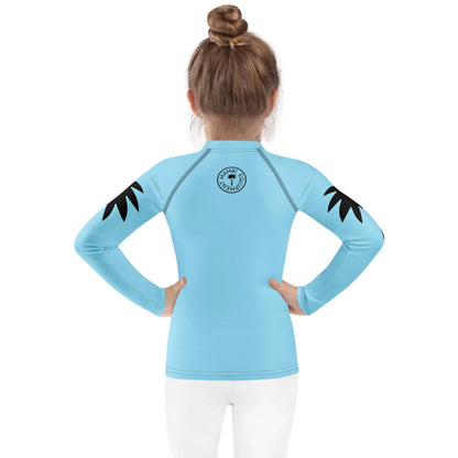 A young child with light brown hair in a bun faces away, wearing a light blue long-sleeve Mambí rash guard with black palm designs on the shoulders and a circular logo on the upper back. The child is also wearing white pants.