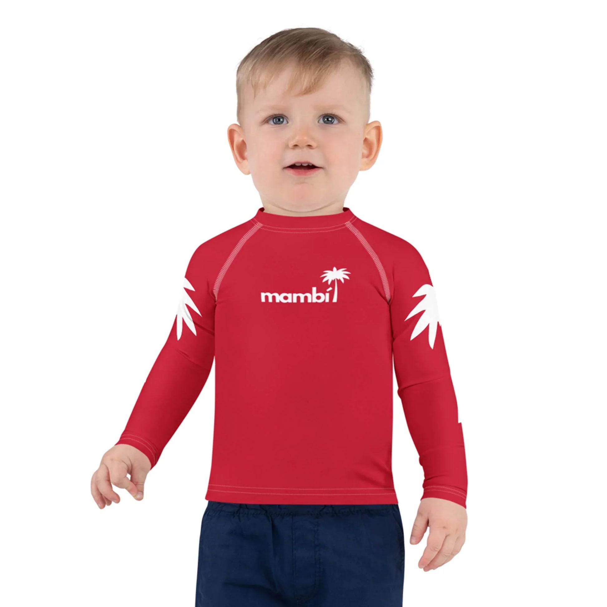 A toddler with short blonde hair wears a red long-sleeve Mambí rash guard featuring white palm designs and the word “mambi” on the chest, paired with dark blue shorts, standing against a plain white background.