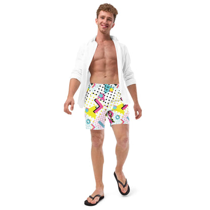 A smiling man in a white open shirt, colorful Mambí patterned shorts, and black flip-flops stands against a white background, looking relaxed and confident.
