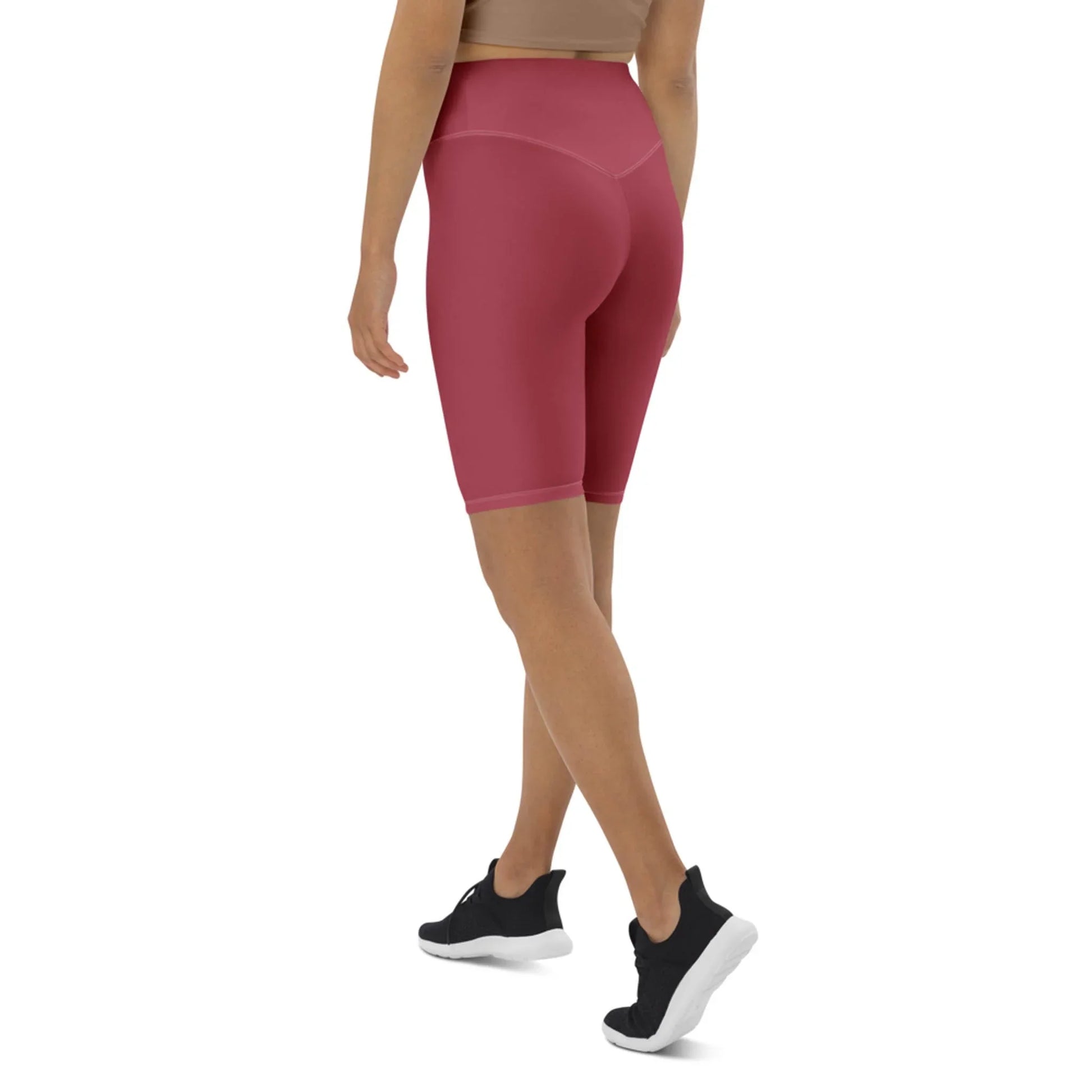 A woman wearing a tan Mambí sports bra, high-waisted pink bike shorts, and black athletic shoes stands with her back turned, walking away on a white background.
