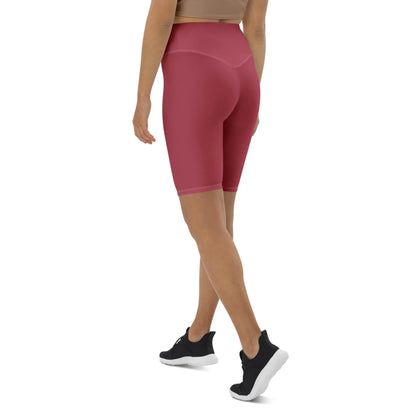 A woman wearing a tan Mambí sports bra, high-waisted pink bike shorts, and black athletic shoes stands with her back turned, walking away on a white background.