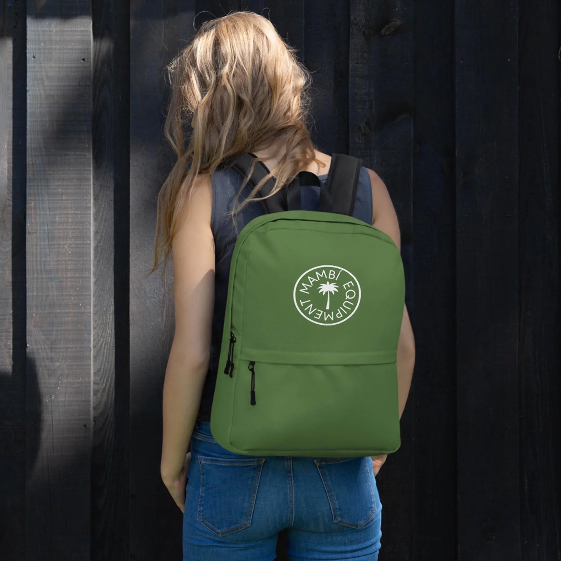 A woman with long, wavy blonde hair, dressed in a black sleeveless top and blue jeans, stands facing a dark wooden fence. She carries the Printful Boulevard East All-Purpose Backpack featuring a circular palm tree logo and hidden back pocket.