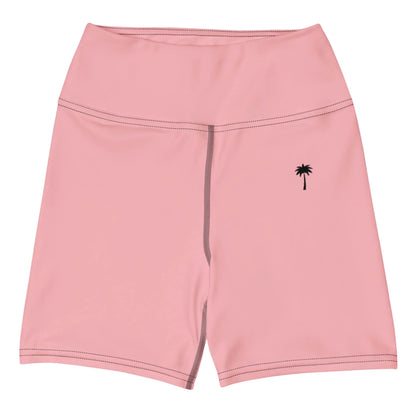 The Brickell Yoga Shorts by Printful are pink, high-waisted shorts with a high waistband, dark stitching, and a small black palm tree graphic on the left leg. Made from four-way stretch fabric. Displayed flat on a white background.