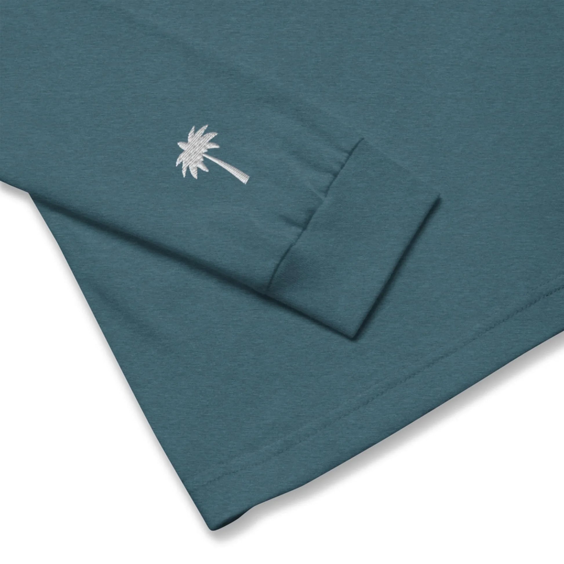 A teal long-sleeve Mambí shirt featuring a small white embroidered palm tree near the cuff of the sleeve.
