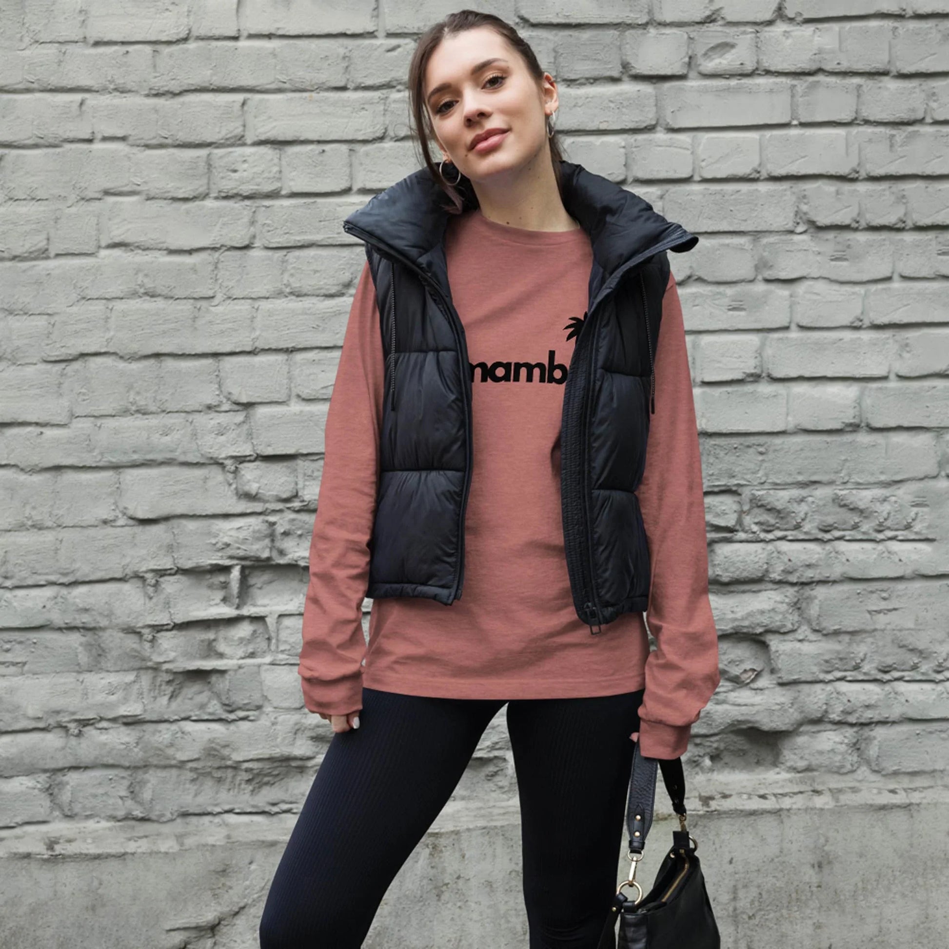 A woman stands in front of a gray brick wall, wearing a pink long-sleeve Mambí shirt with black text, a black puffer vest, black pants, and holding a black handbag. She has her hair tied back and is looking at the camera.