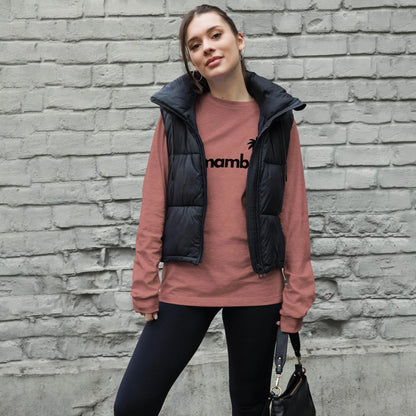 A woman stands in front of a gray brick wall, wearing a pink long-sleeve Mambí shirt with black text, a black puffer vest, black pants, and holding a black handbag. She has her hair tied back and is looking at the camera.