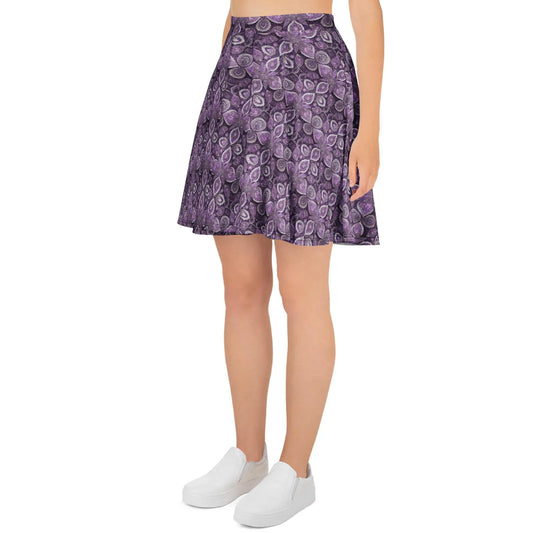 A woman models the Printful Candelaria Skater Skirt, featuring a high waist, purple pattern, and elastic waistband. She pairs it with white slip-on shoes and a cropped top; only her lower body shows against a white background.