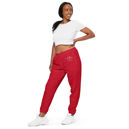 A woman in a white Mambí crop top and red jogger pants stands sideways, looking to her left and smiling. She poses with one hand on her hip, wearing white sneakers against a plain white background.