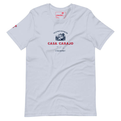White t-shirt with a small illustration of a house and palm tree on the chest, featuring “The Ultimate Destination CASA CARAJO If you can’t find it, you can’t do it.” in blue and red. Includes a Mambí-inspired red palm icon on one sleeve.