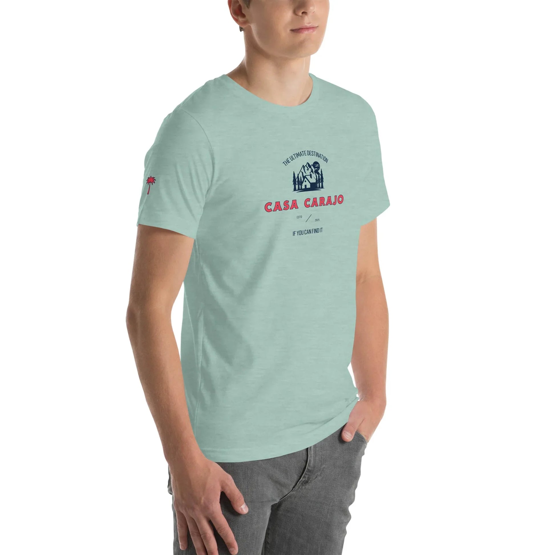 A person wearing a light green Mambí t-shirt with Casa Carajo and an illustrated design on the chest, standing with one hand in a gray pocket against a white background.