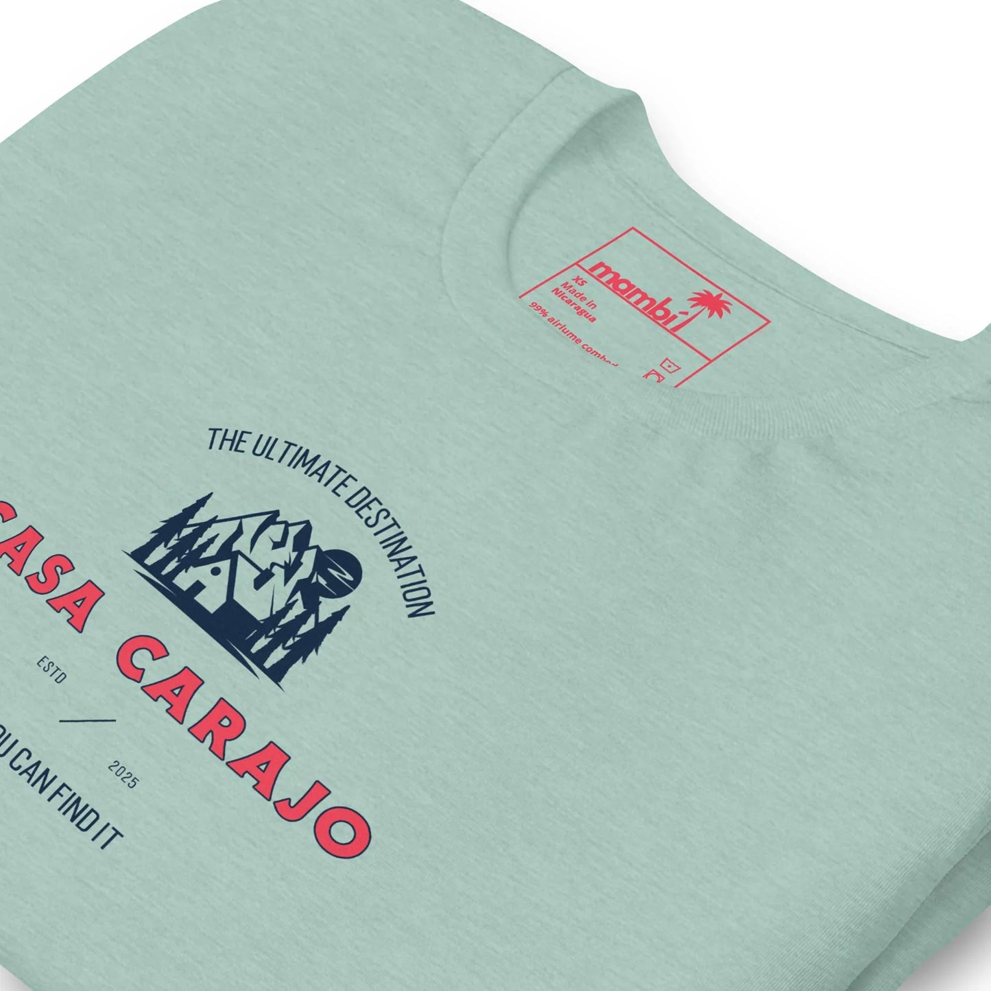 A light green t-shirt with a printed design featuring a mountain, trees, and the text “The Ultimate Destination CASA CARAJO” in bold red and blue letters. Mambí-inspired small text and graphics appear below the main design.