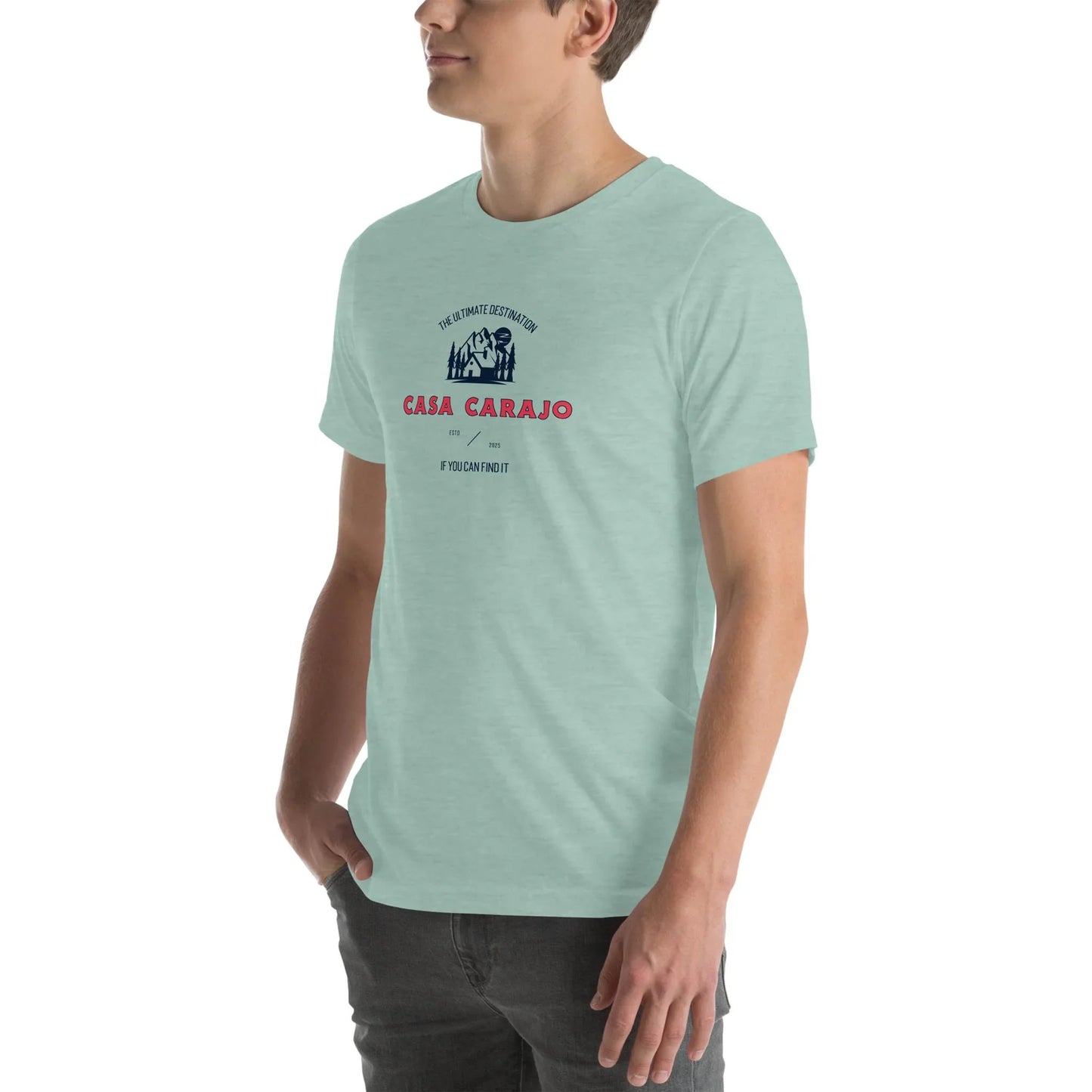 A person in a light mint-green Mambí t-shirt with Casa Carajo and a small graphic on the chest stands with one hand in their pocket against a plain white background.