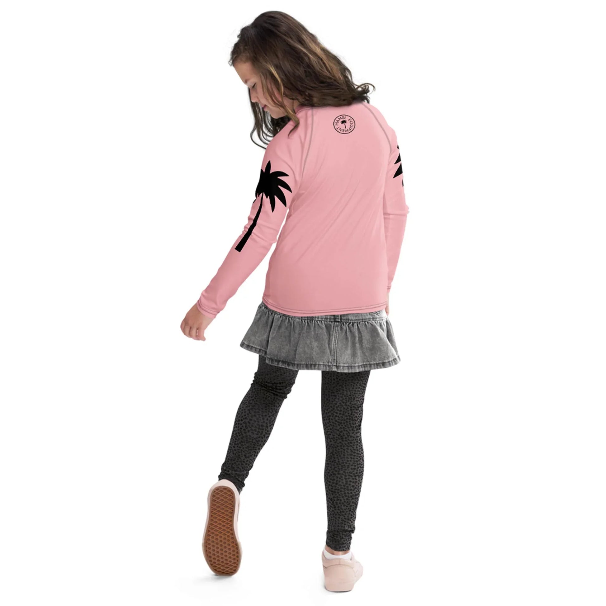 A young girl wearing a pink long-sleeve Mambí shirt with black palm tree designs, a gray ruffled skirt, and patterned leggings walks forward while looking down over her shoulder.