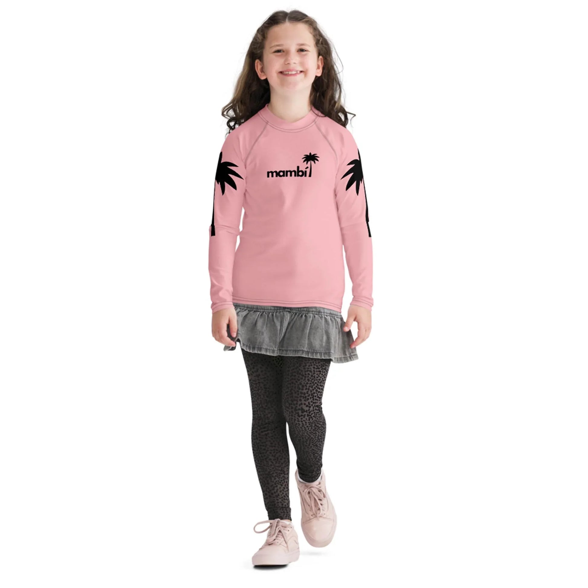 A smiling young girl with long brown hair wears a pink long-sleeve shirt featuring Mambí text and black palm tree graphics, a gray skirt, patterned leggings, and pink shoes as she walks forward.