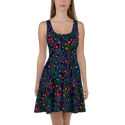 A woman wears the Coco Skater Dress by Printful, featuring a sleeveless design, flared skirt, and vibrant floral pattern in red, orange, yellow, pink, and green. Made from soft fabric, she stands facing forward against a white background.