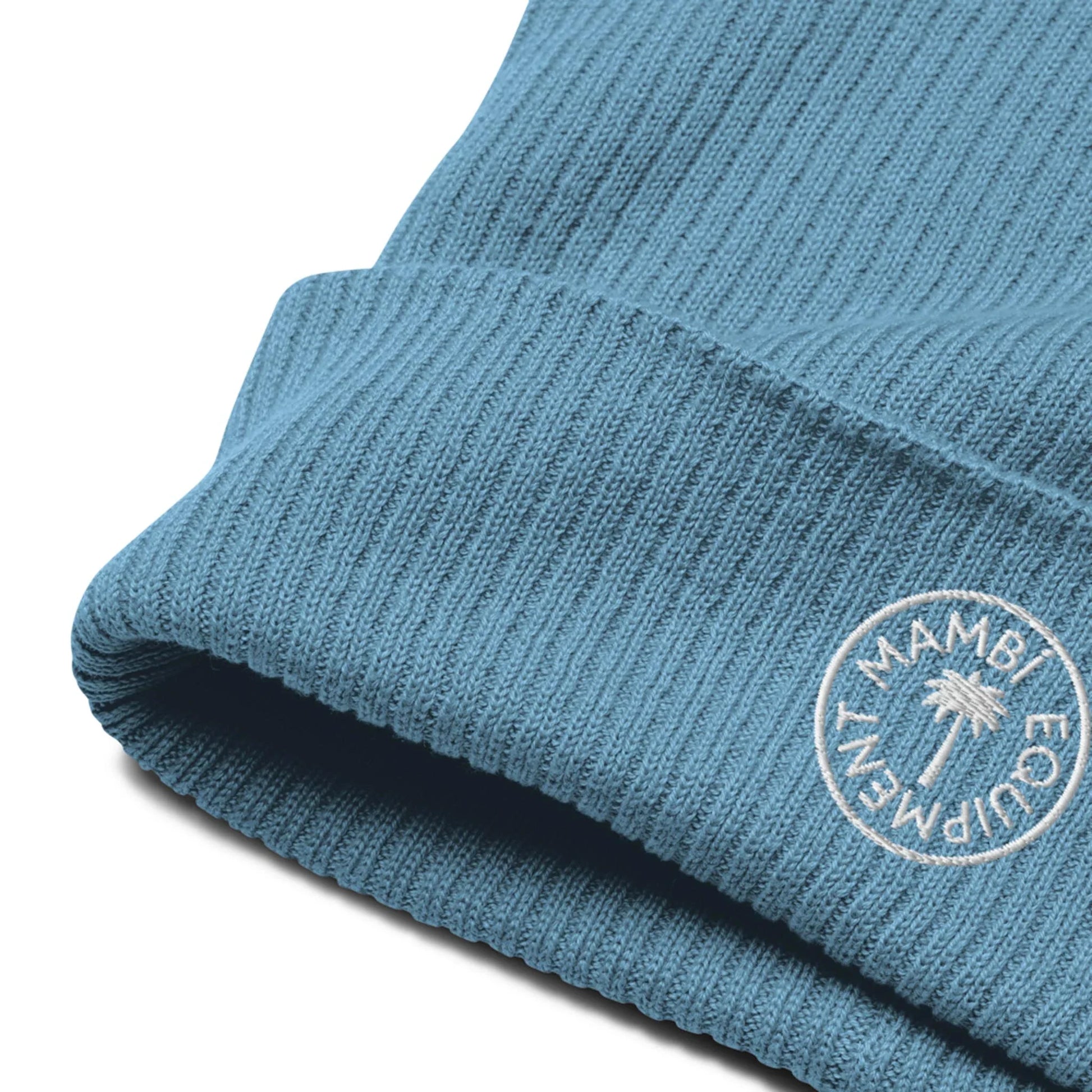Close-up of a blue ribbed knit beanie with a folded brim. The brim features an embroidered white circular Mambí logo and a small palm tree illustration in the center.