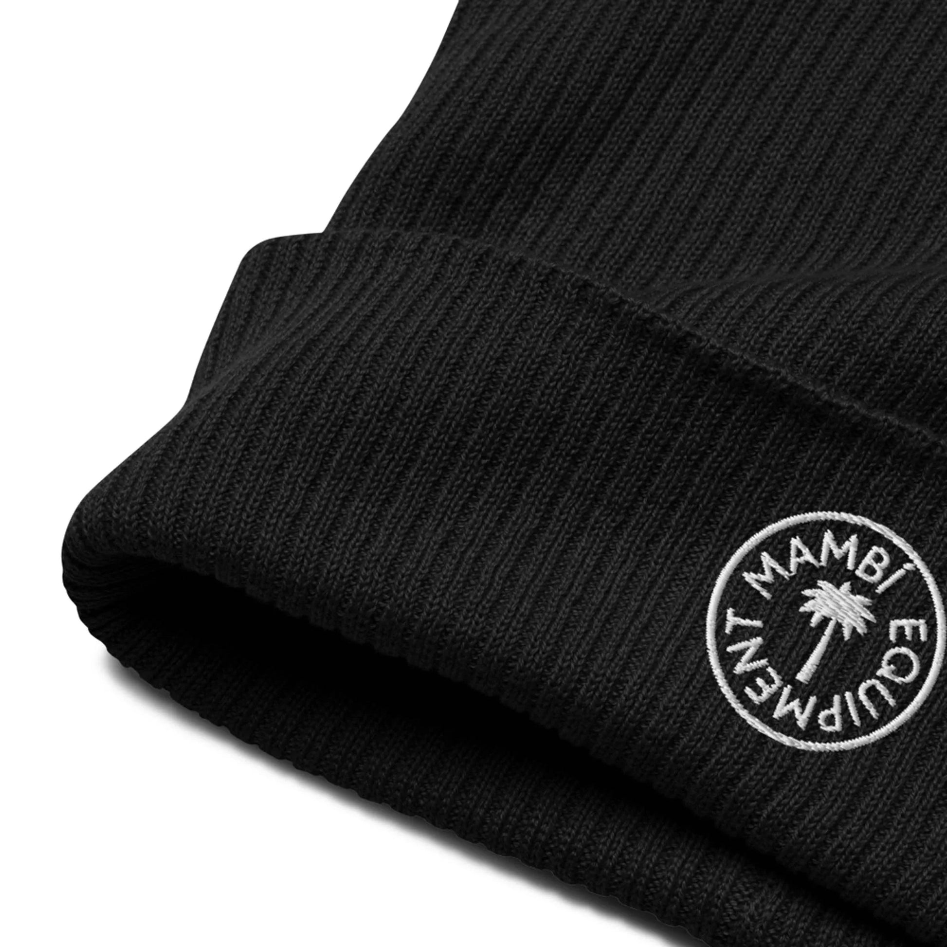 Close-up of a black ribbed knit beanie with a folded brim, featuring a round white embroidered patch that reads Mambí Equipment and includes a small palm tree graphic.