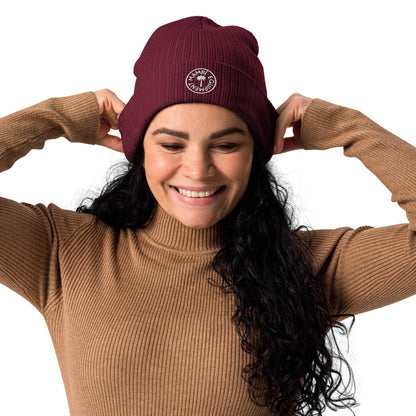 Smiling woman with long curly dark hair wears a ribbed brown sweater and a maroon Mambí beanie, adjusting the hat with both hands. The beanie features a small white circular logo on the front. The background is white.