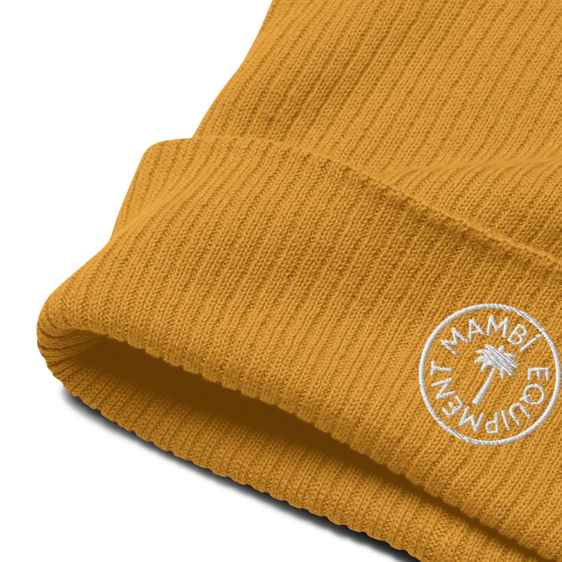 Close-up of a mustard yellow ribbed knit beanie with a folded brim, featuring a white circular embroidered Mambí logo that includes the text “MAMBI EQUIPMENT” and a palm tree design.