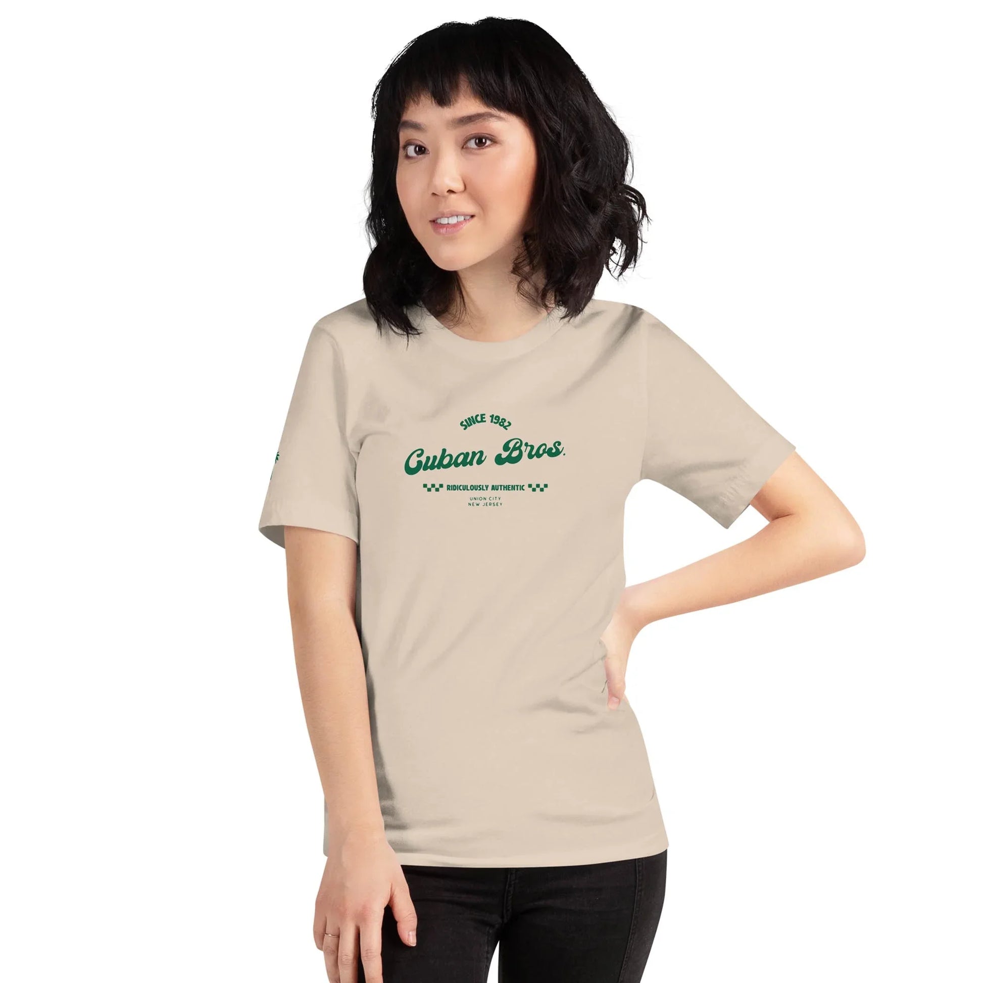 A woman with shoulder-length dark hair wears the Printful Cuban Bros Graphic Tee | Unisex from the Neighborhood Collection, featuring Since 1998 Cuban Bros in green, standing with one hand on her hip and smiling at the camera.