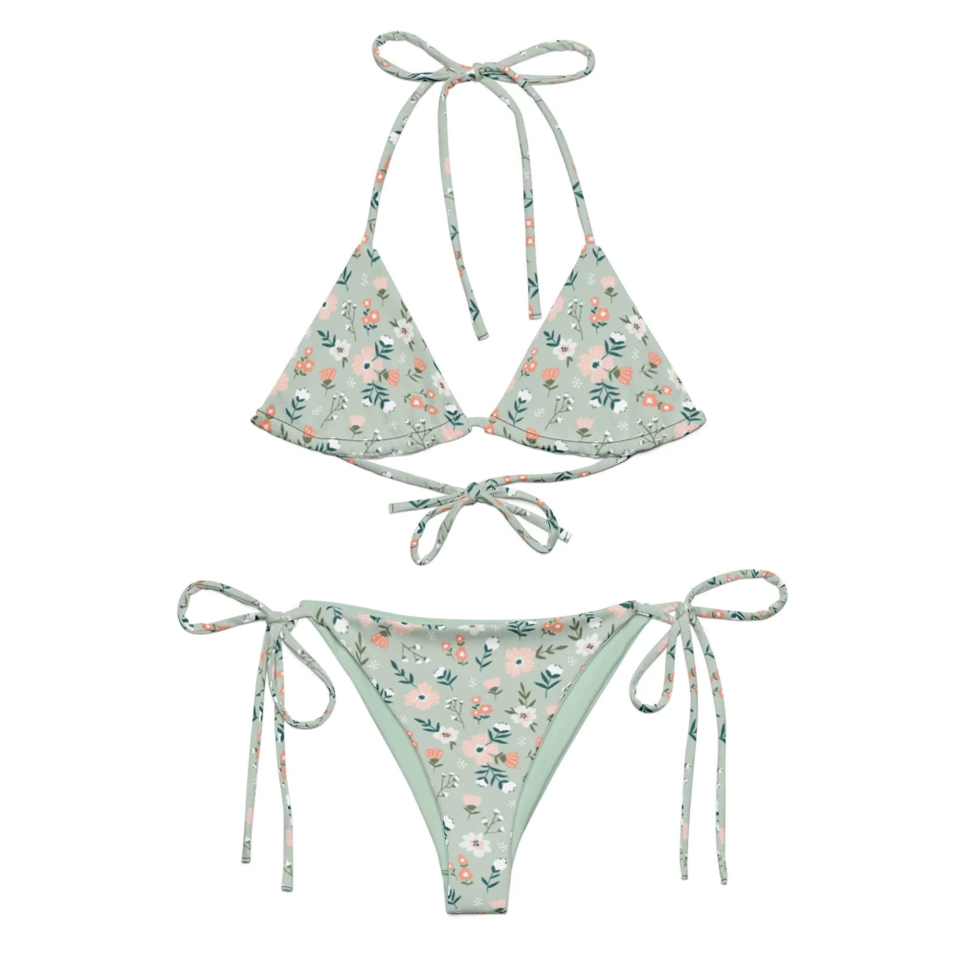 The Daniella String Bikini Swimsuit by Printful features a light green floral print with pink and white flowers, is made from recycled polyester with UPF 50+, and has a triangle top with neck/back ties and adjustable side-tie bottoms.