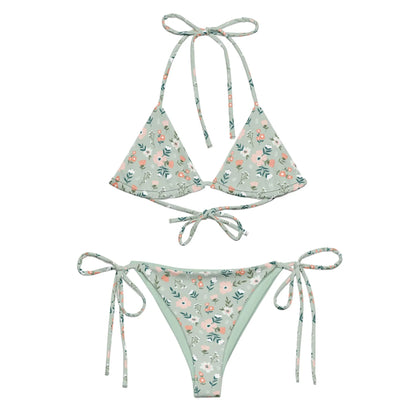 The Daniella String Bikini Swimsuit by Printful features a light green floral print with pink and white flowers, is made from recycled polyester with UPF 50+, and has a triangle top with neck/back ties and adjustable side-tie bottoms.