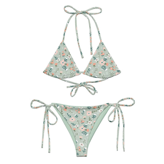 The Daniella String Bikini Swimsuit by Printful features a light green floral print with pink and white flowers, is made from recycled polyester with UPF 50+, and has a triangle top with neck/back ties and adjustable side-tie bottoms.